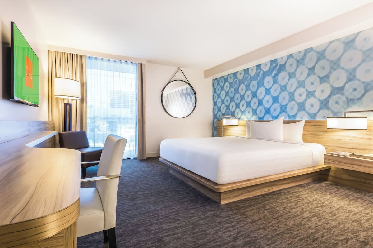 A modern hotel room with a large bed, a desk, and a chair. There is a TV mounted on the wall, a lamp, and a window with curtains.