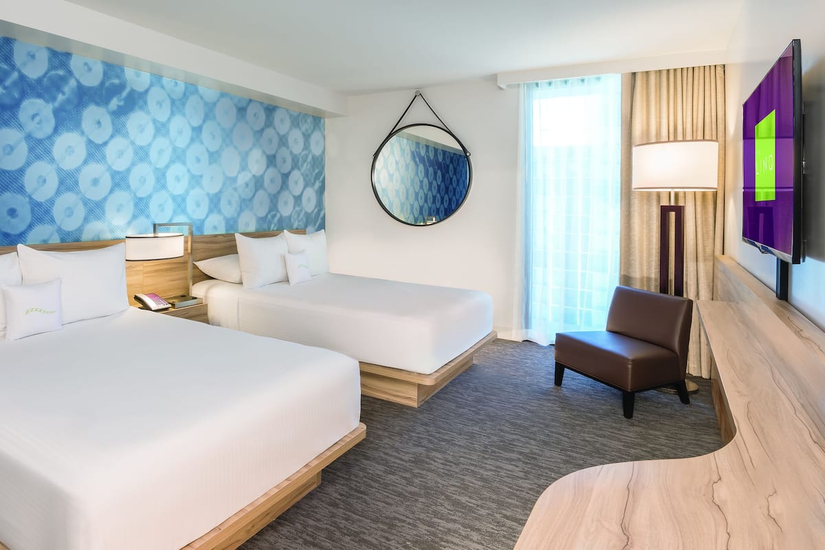 A hotel room with two beds, a desk, a chair, a TV, and a patterned wall.