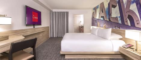 Habitación de hotel con una cama grande, un escritorio, una silla, un televisor y un baño visible a través de una puerta de vidrio.