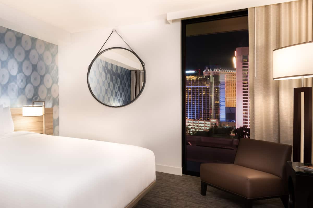 A hotel room with a large bed, a round mirror, a chair, and a view of a cityscape at night.
