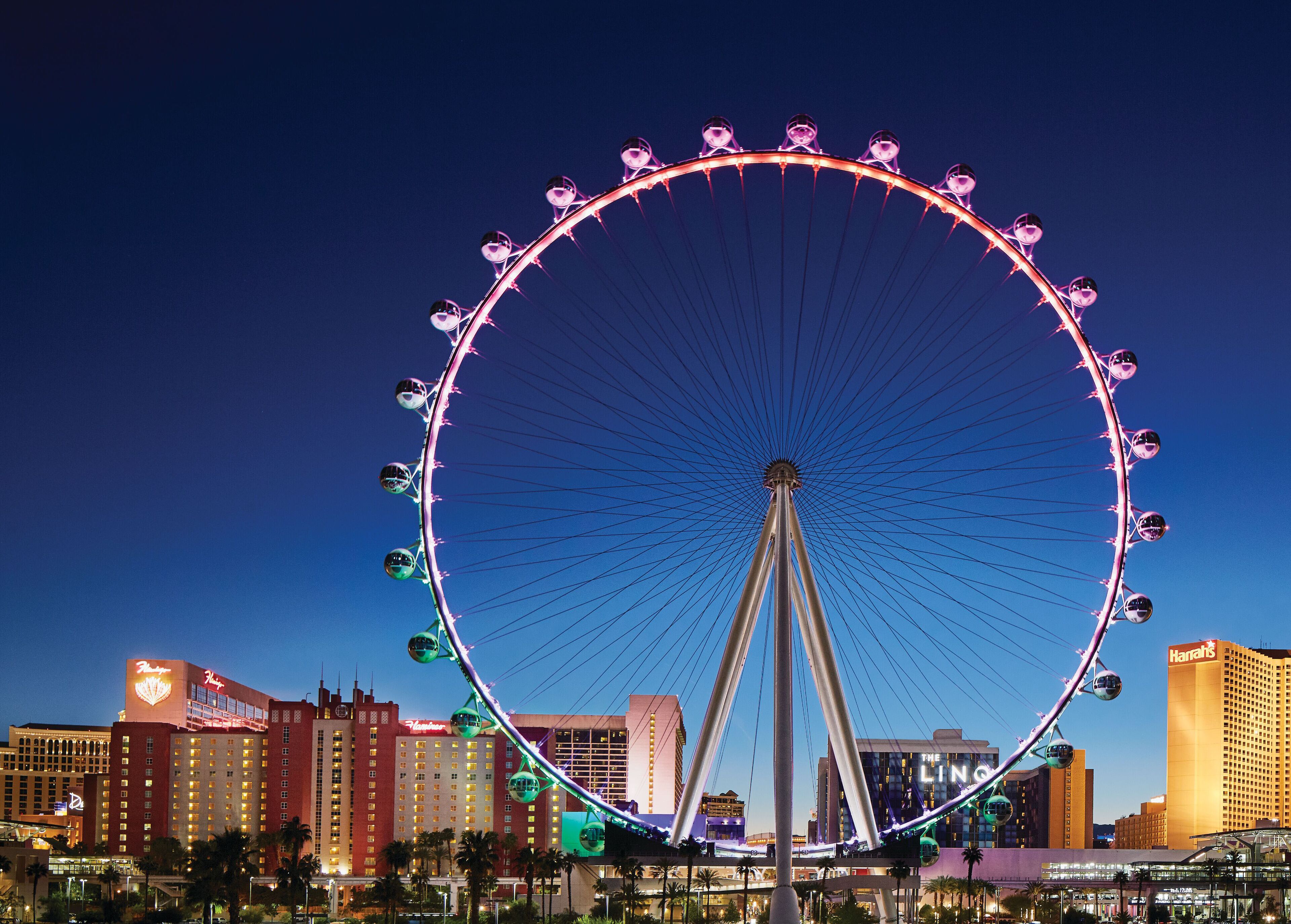 A large Ferris wheel with multiple cabins, surrounded by a city skyline with various buildings, including a prominent hotel with a neon sign.