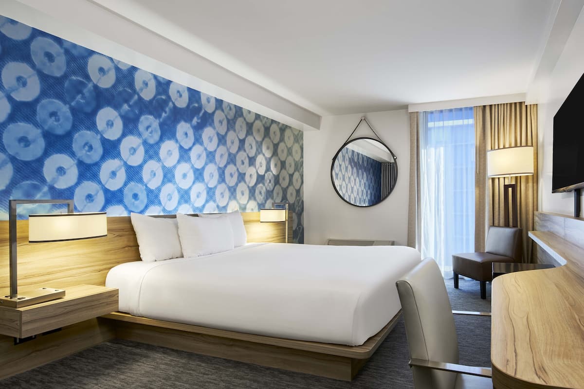 A modern hotel room with a large bed, a desk, a chair, a TV, and a patterned wall.