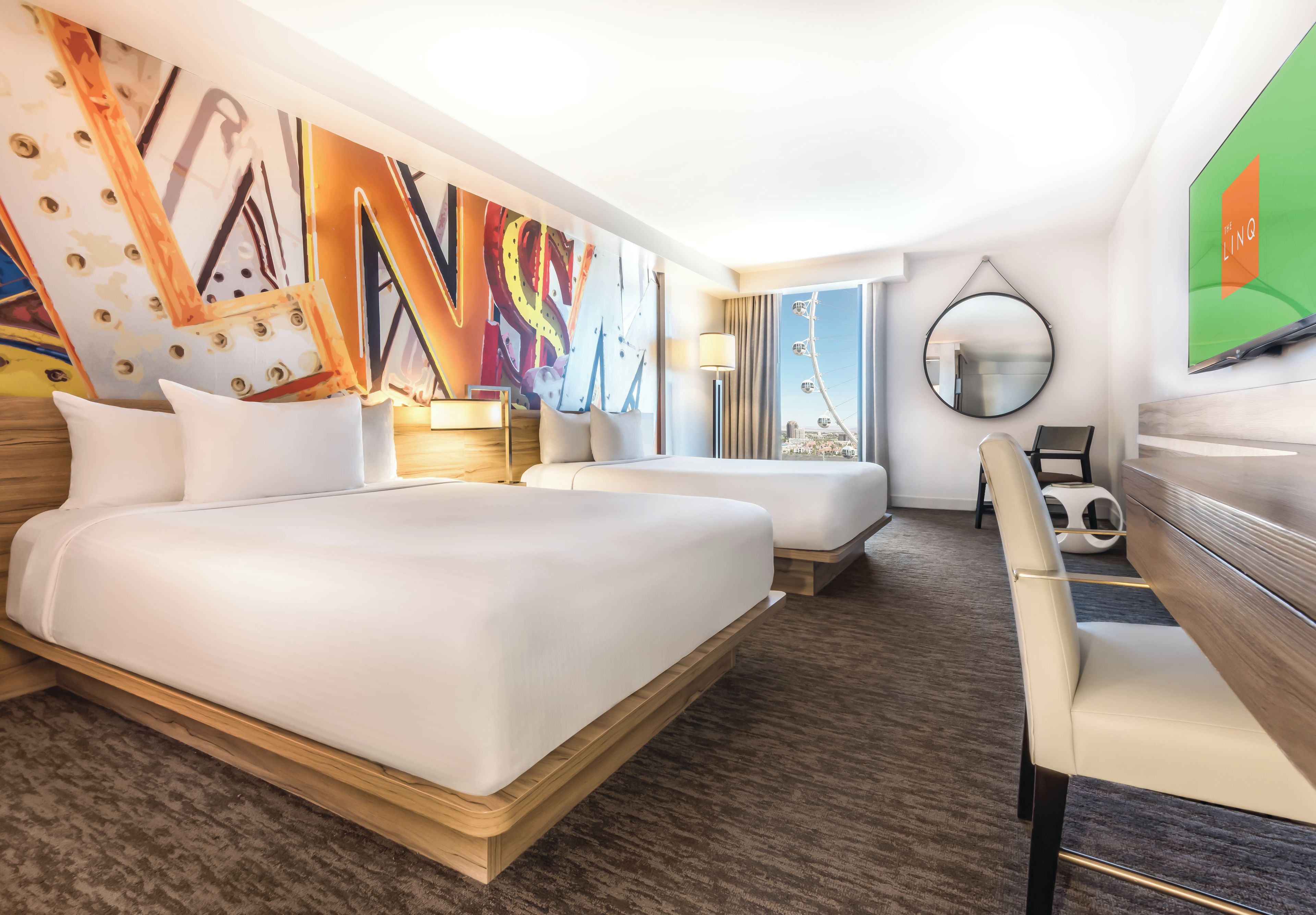 Ein Hotelzimmer mit zwei Betten, einem Schreibtisch, einem Stuhl, einem großen Spiegel und einer Wand mit abstrakten Kunstwerken.