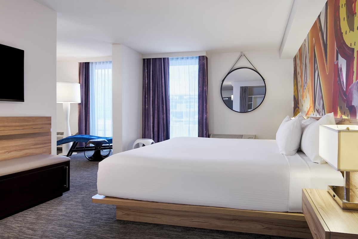 A hotel room with a large bed, a desk, a chair, a TV, and a painting on the wall.