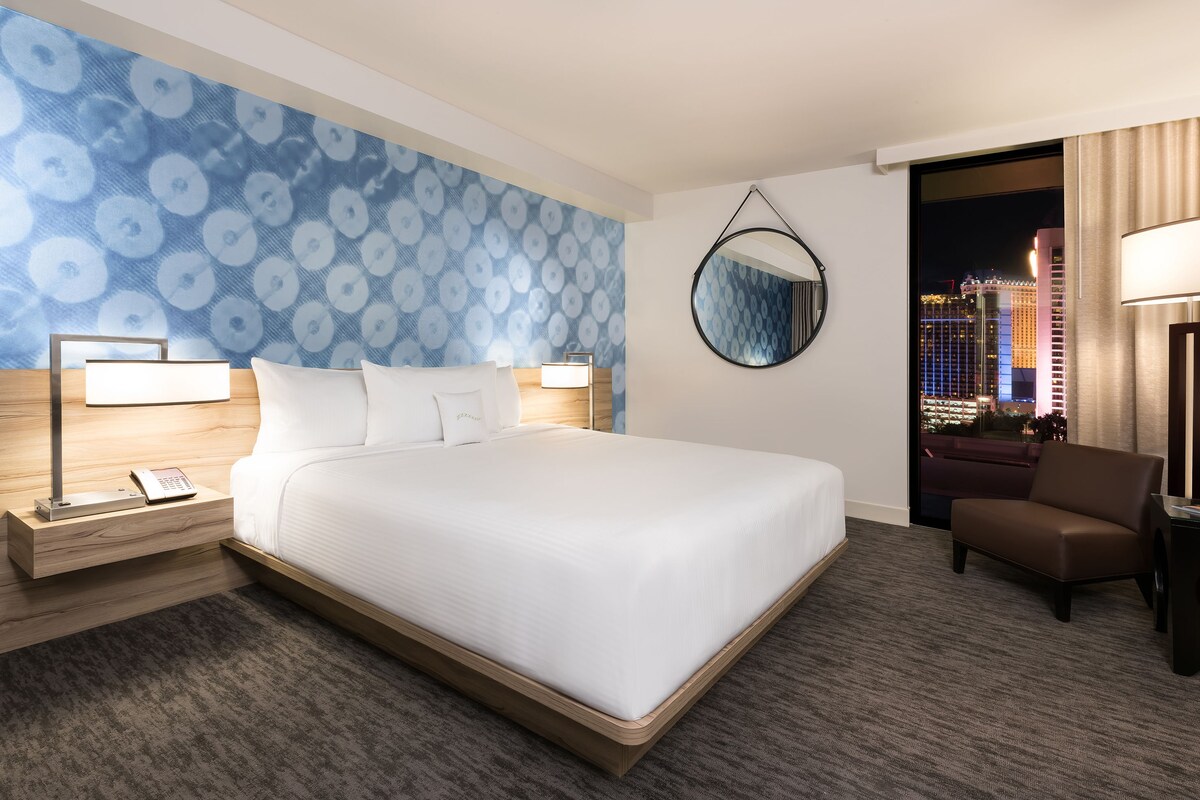 A hotel room with a large bed, bedside lamps, a nightstand, a chair, a mirror, and a view of a cityscape.