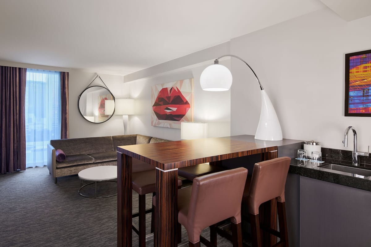 A modern hotel room with a sofa, a small round table, a bar area with stools, a TV, and a large window with curtains.