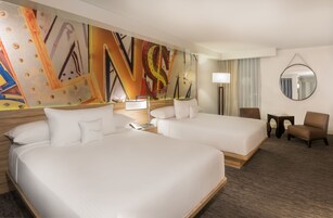 Quarto de hotel com duas camas, uma grande pintura abstrata na parede, um espelho redondo e uma pequena mesa com abajur.