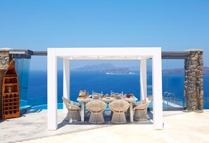 An outdoor dining area with a table set for four, overlooking a clear blue sea and distant islands.