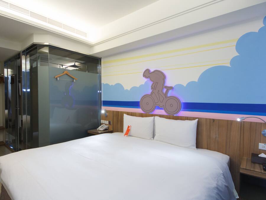 Ein Hotelzimmer mit einem großen Bett, einer gläsernen Dusche, einem Wandgemälde mit einem Fahrrad und einem hölzernen Kopfteil.