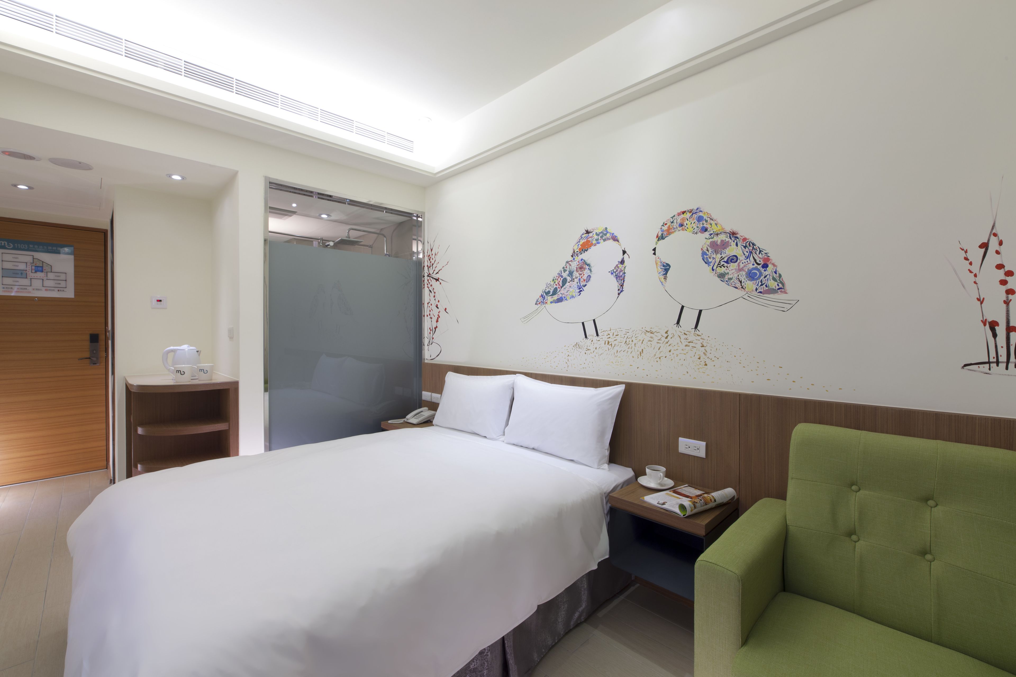A hotel room with a large bed, a green armchair, a nightstand with a phone and a cup, and a wall with bird artwork.