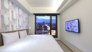 Habitación de hotel con una cama grande, vista a la ciudad a través de una puerta corrediza, televisor montado en la pared y un papel tapiz estampado.