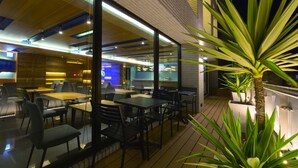 Un área de comedor al aire libre con mesas y sillas, un muro de vidrio y una planta en maceta.
