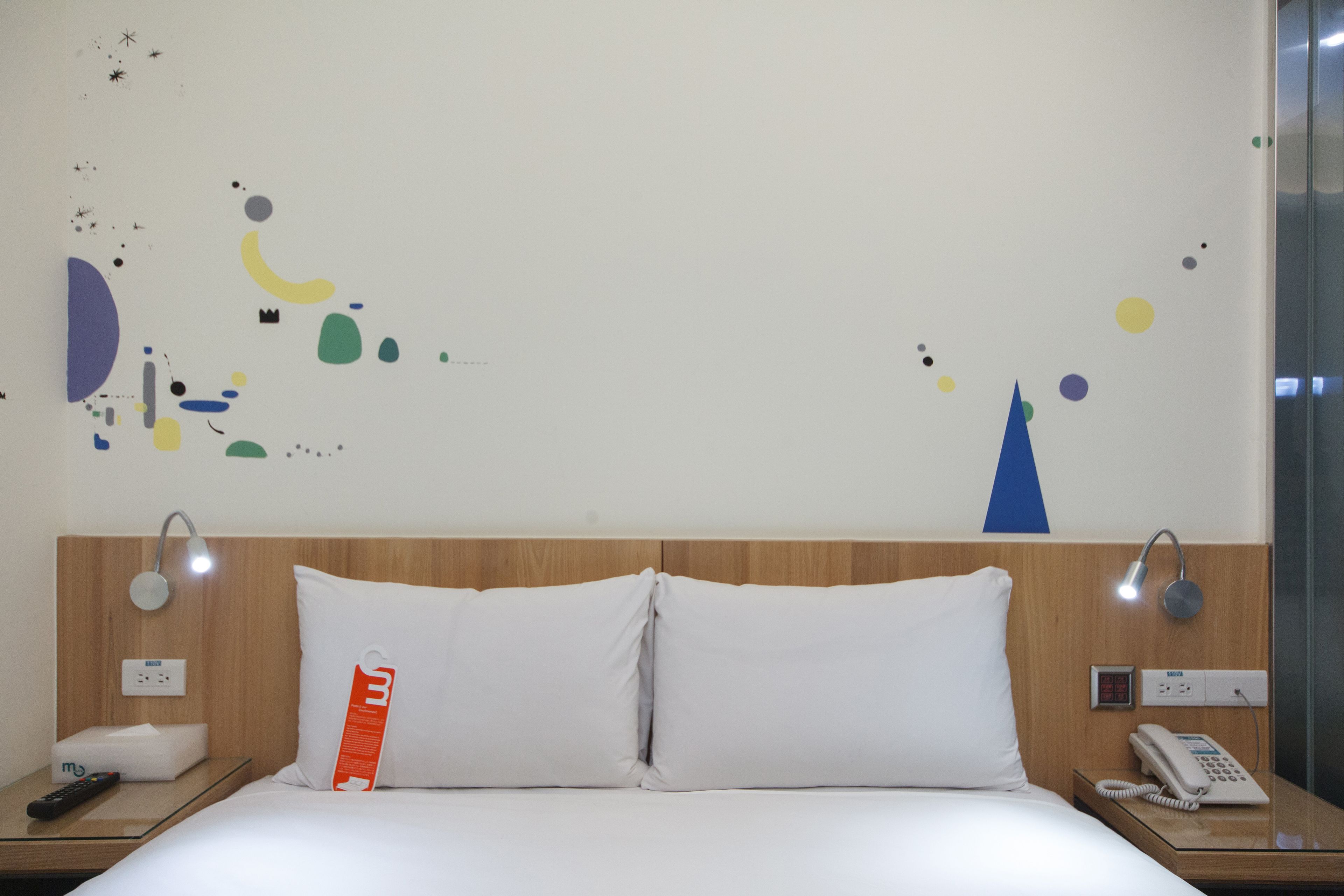 Quarto de hotel com cama, mesa de cabeceira, telefone e parede decorada com arte abstrata.