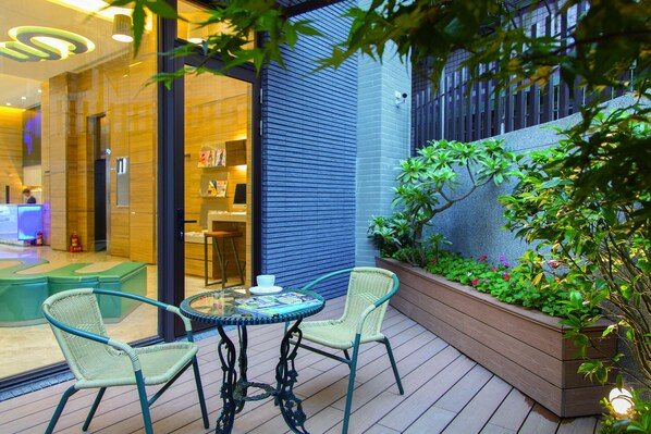 Un área de estar al aire libre con sillas de mimbre y una mesa de vidrio, rodeada por una jardinera con follaje y flores.