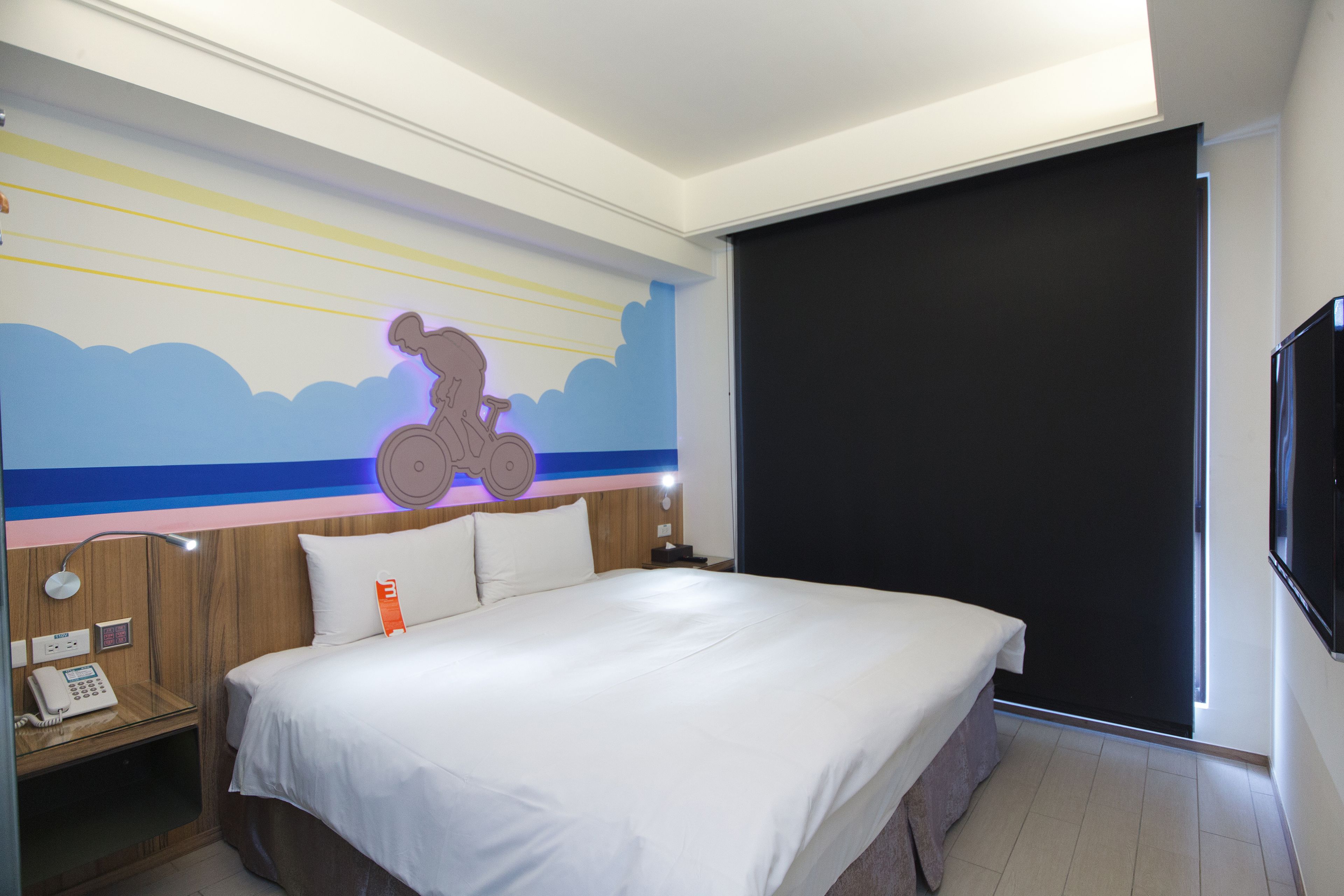 A hotel room with a large bed, a wall-mounted TV, a bedside table with a phone, and a mural of a cyclist.