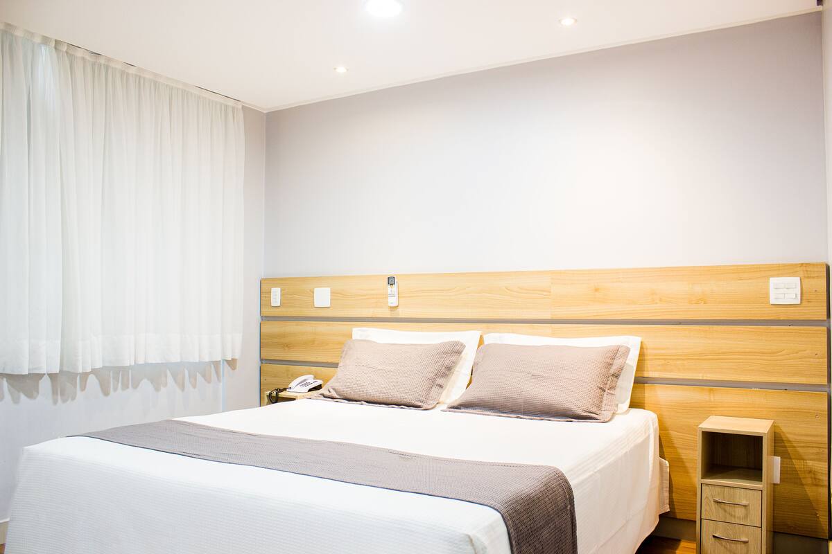 A neatly made bed with a wooden headboard, a bedside table with a lamp, and a small drawer unit.