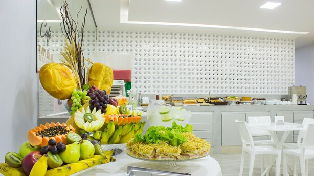 A buffet table with a variety of fruits and a layered salad, white chairs, and a modern kitchen in the background. - Imperial Plaza Hotel (Samambaia)