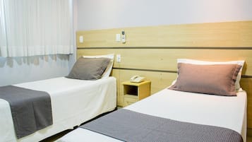 A hotel room with two single beds, each with a pillow and a blanket. There is a wooden headboard, a bedside table with a phone, and a power outlet on the wall.