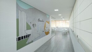 A corridor with a mural, white walls, and a long table with chairs. - Imperial Plaza Hotel (Samambaia)