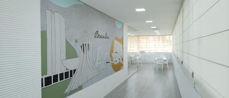 Um corredor com um mural, paredes brancas e uma mesa comprida com cadeiras.