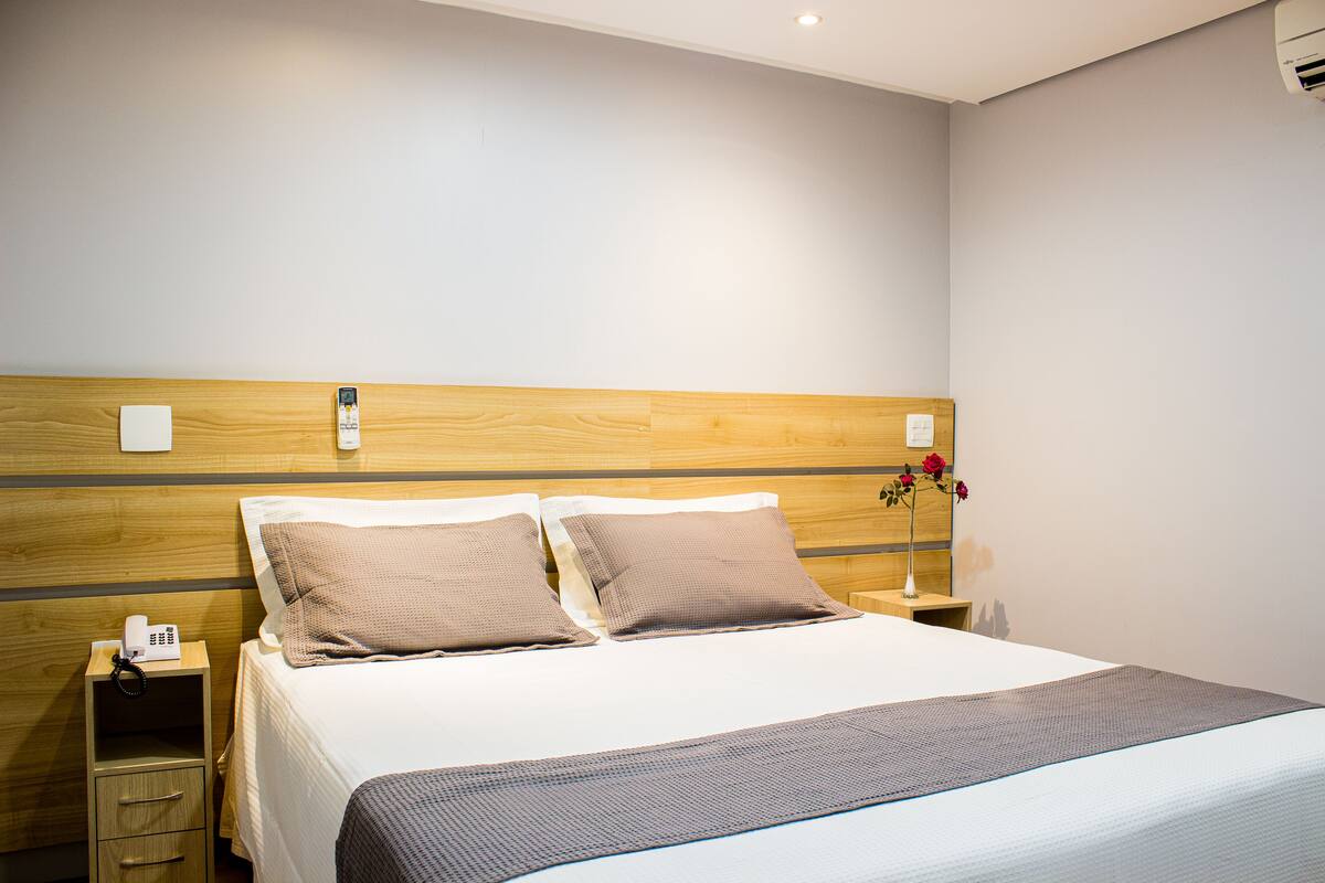 A neatly made bed with two pillows, a bedside table with a phone, and a vase with flowers on a nightstand.