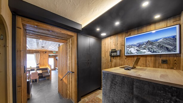 A modern interior with a wooden reception desk, a mounted TV displaying a mountainous landscape, and a view into a dining area. - Hotel Gasthof Siggen (Neukirchen am Grossvenediger)