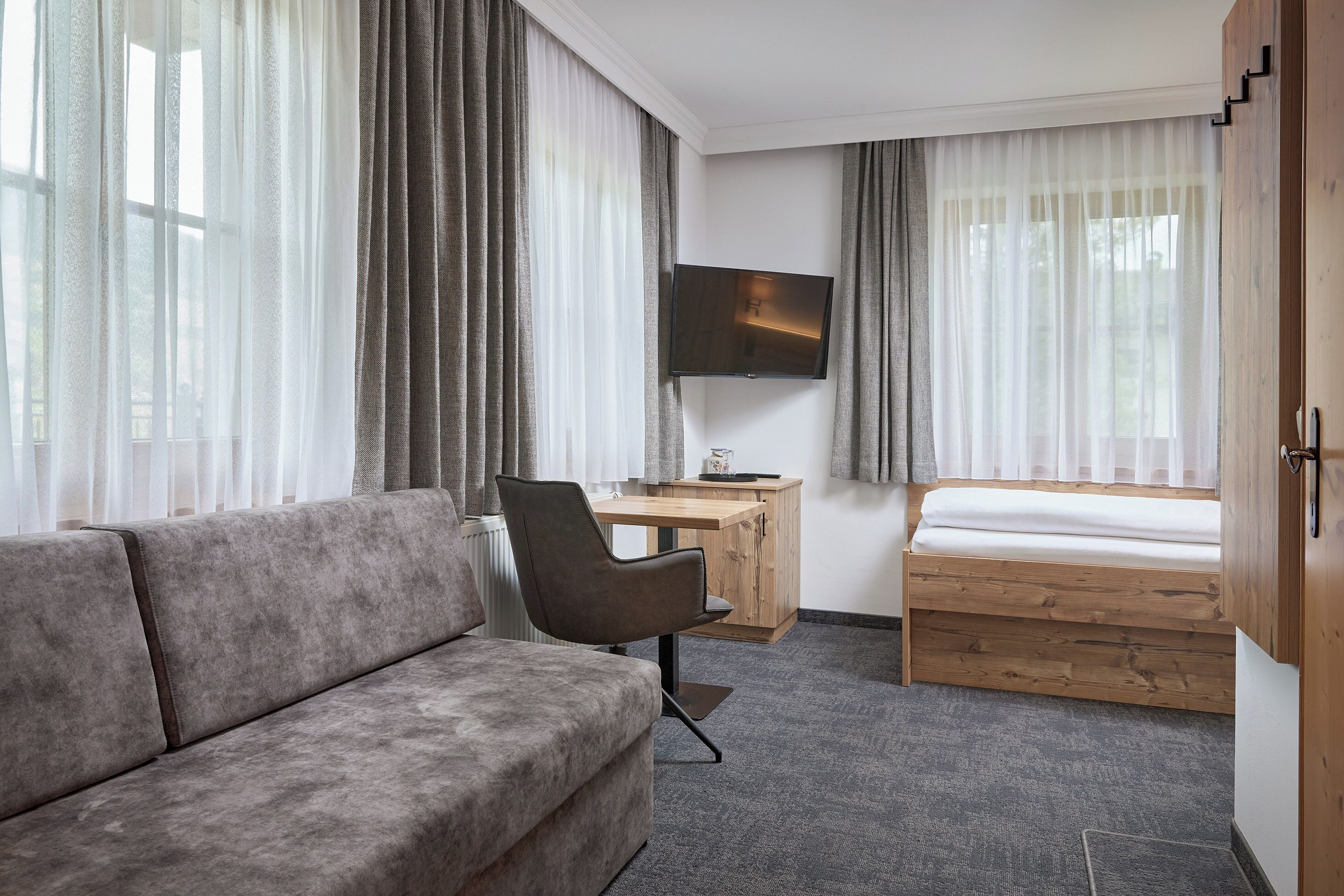 A modern hotel room with a sofa, a wooden desk, a bed, and a TV mounted on the wall.