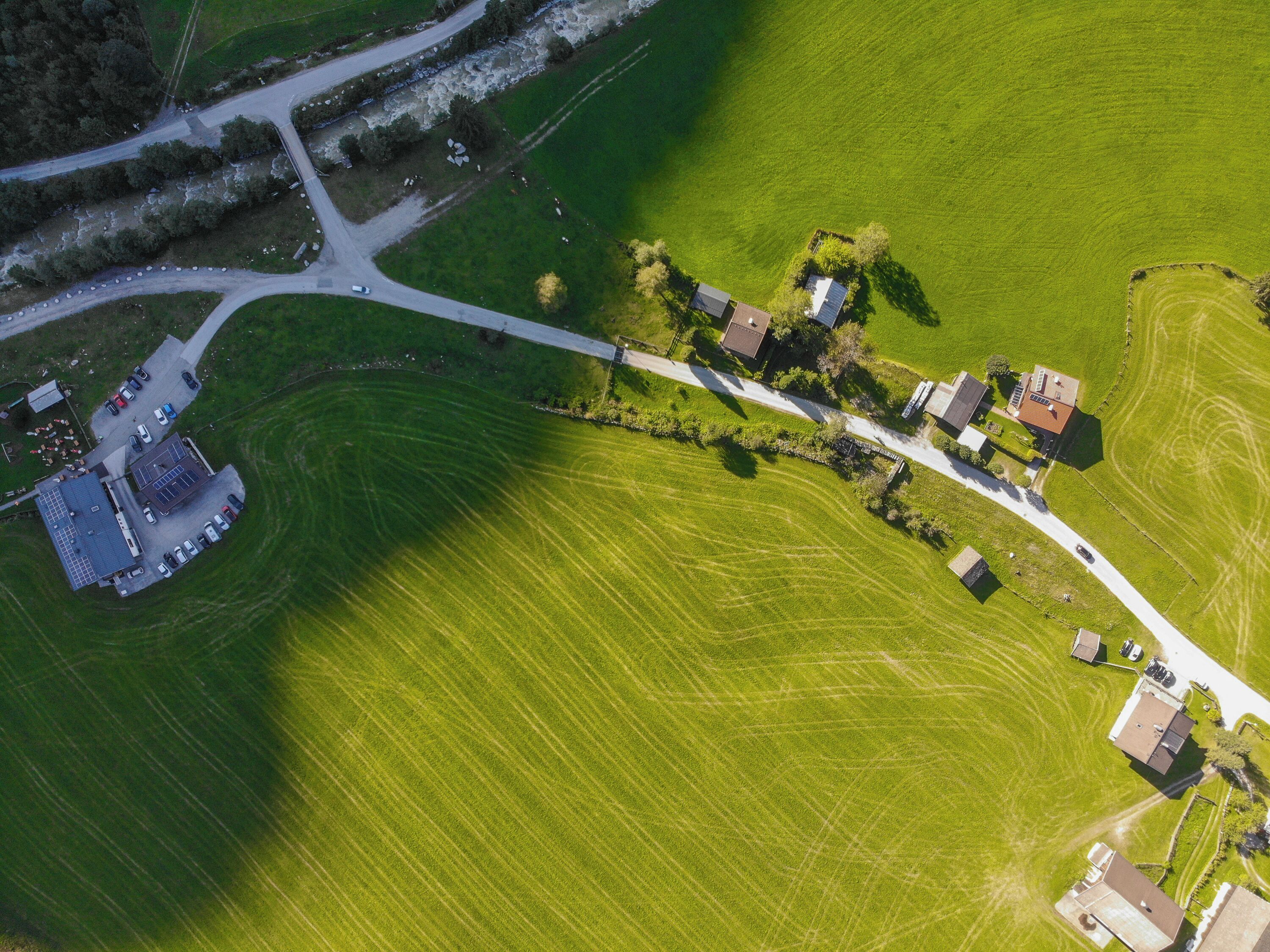 An aerial view of a rural area with a road, green fields, and several buildings including a large structure with a blue roof and a smaller building with a red roof.