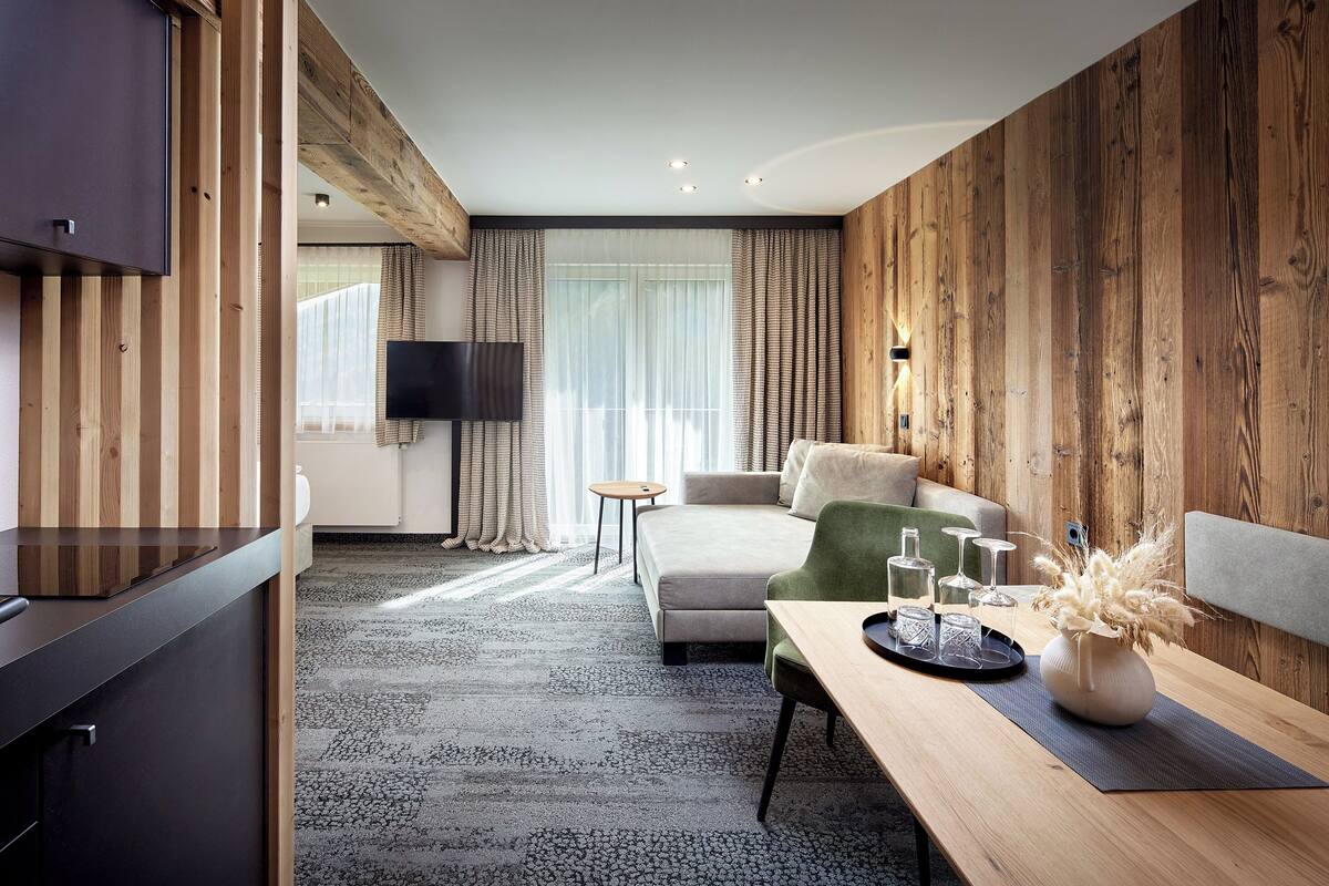 A modern hotel room with a wooden accent wall, a sofa, a dining table with a vase, and a TV mounted on the wall.