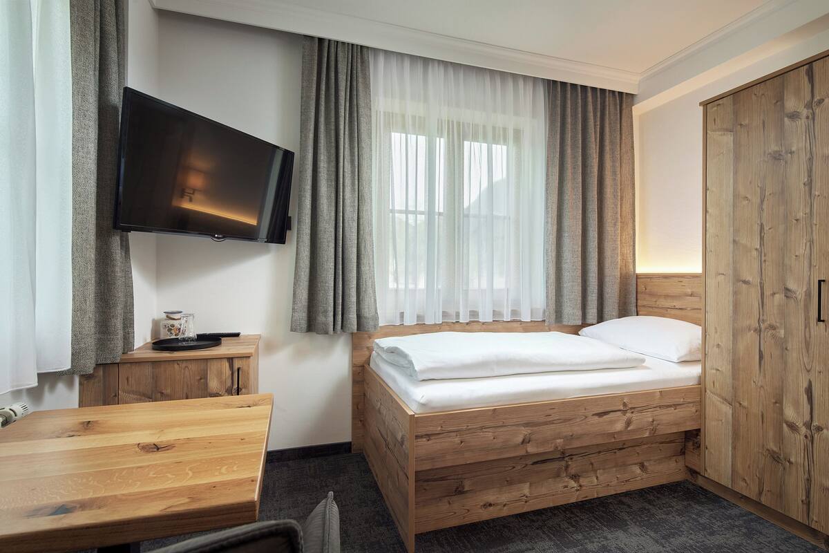 A hotel room with a wooden bed, a flat-screen TV mounted on the wall, a small wooden table, and a window with sheer curtains.
