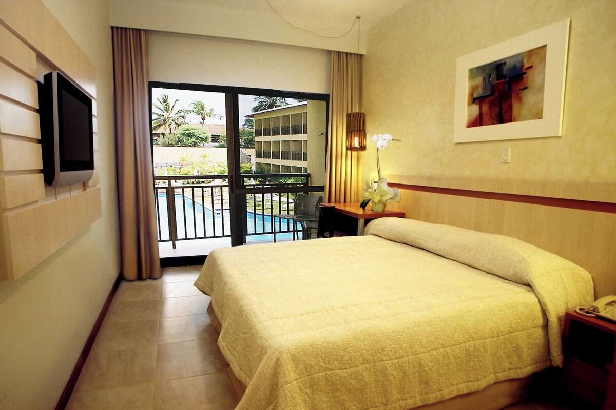 A hotel room with a bed, a TV mounted on the wall, a desk with a lamp, and a view of a pool and building outside.