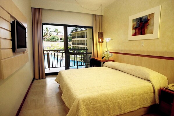 A hotel room with a bed, a TV mounted on the wall, a desk with a lamp, and a view of a pool and building outside. - Catussaba Business (Salvador)