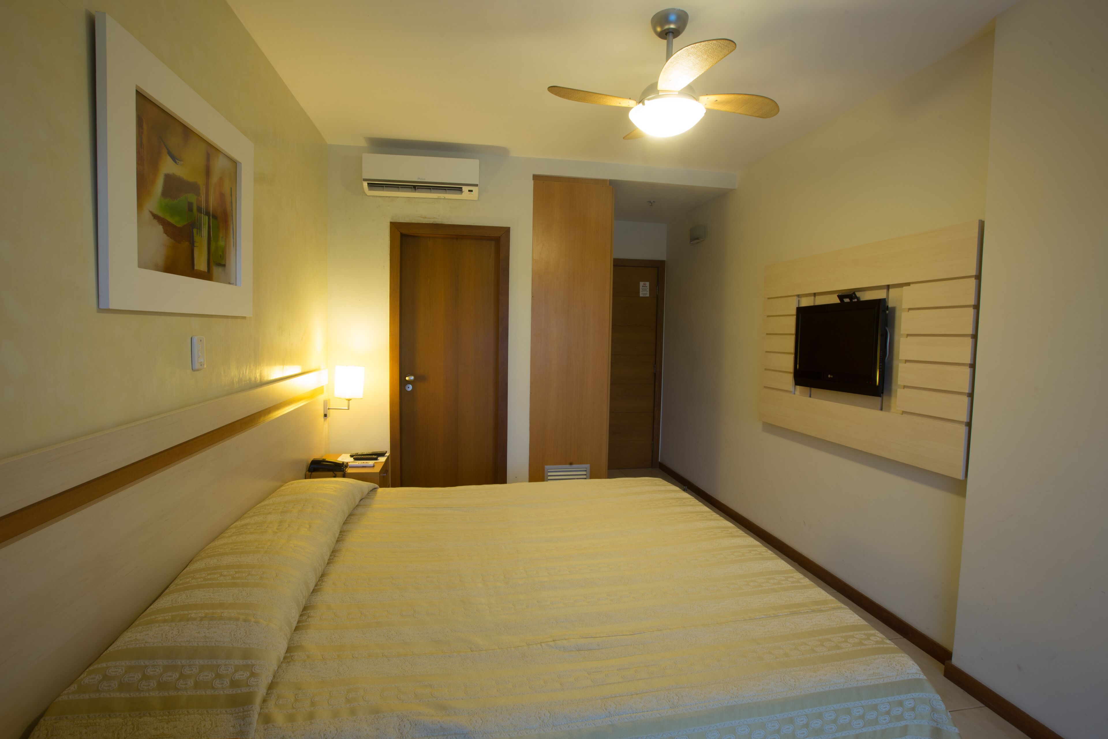 A hotel room with a bed, a ceiling fan, a mounted TV, and a framed picture on the wall.