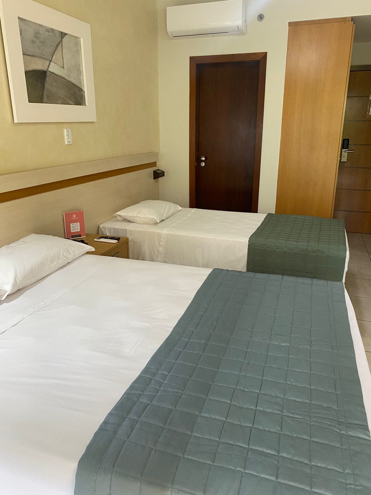 Habitación de hotel con dos camas, una puerta de madera, un equipo de aire acondicionado y un cuadro en la pared.