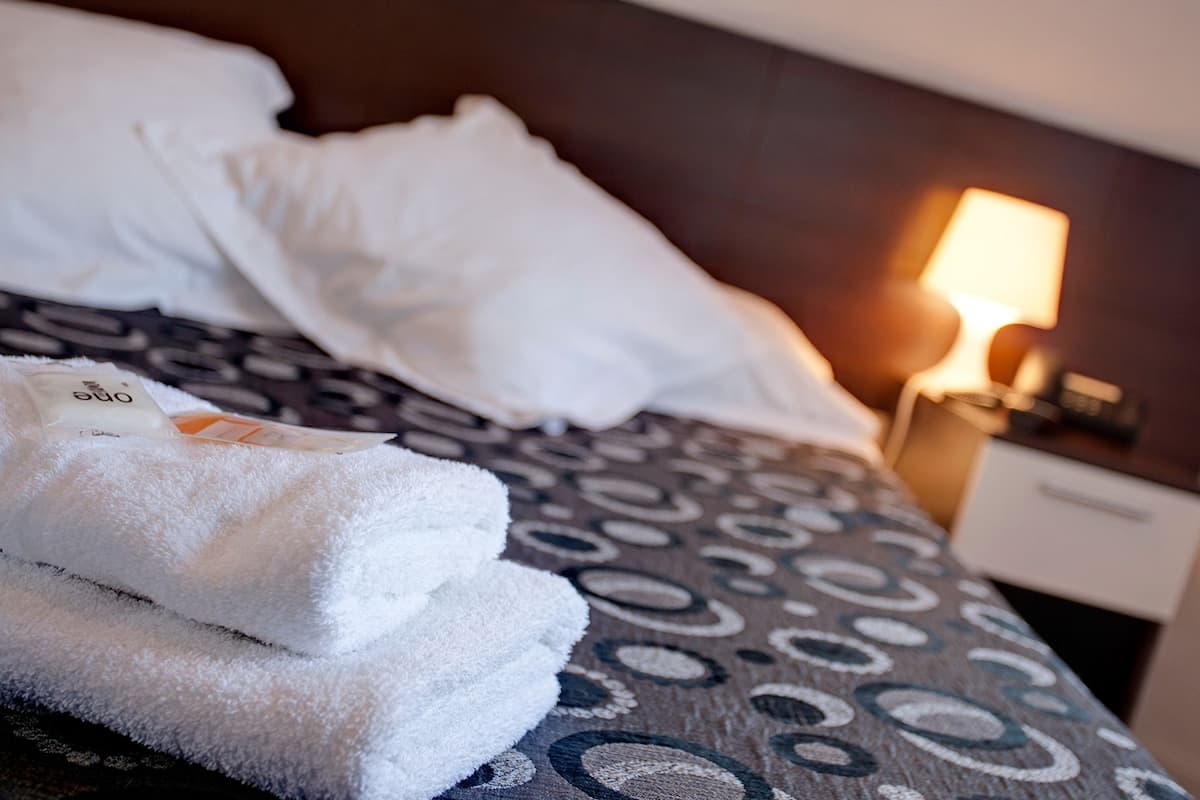 A neatly made bed with white pillows and a patterned bedspread. A white towel is folded on the bed.
