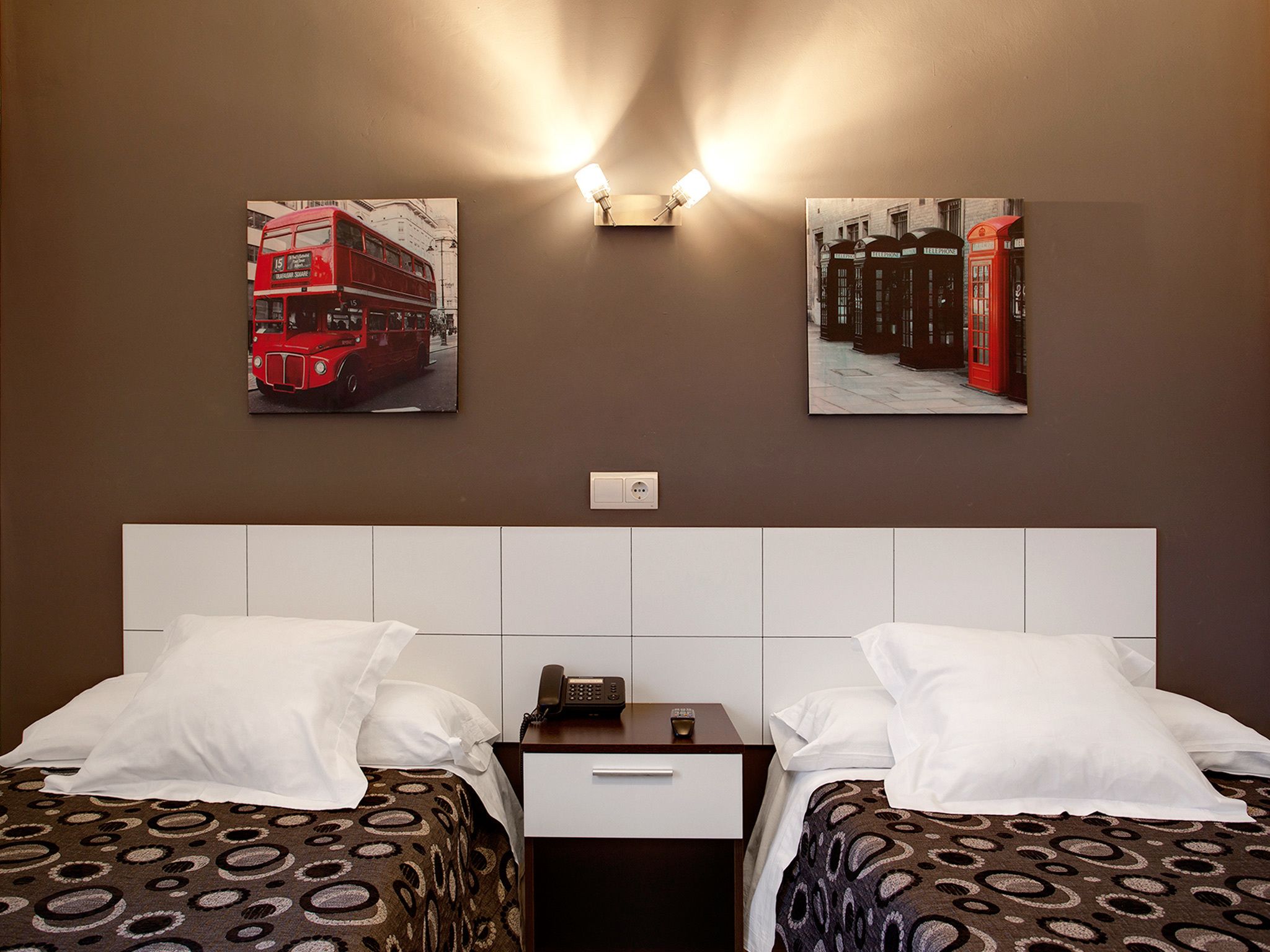 A hotel room with two beds, a nightstand, and a wall-mounted light.