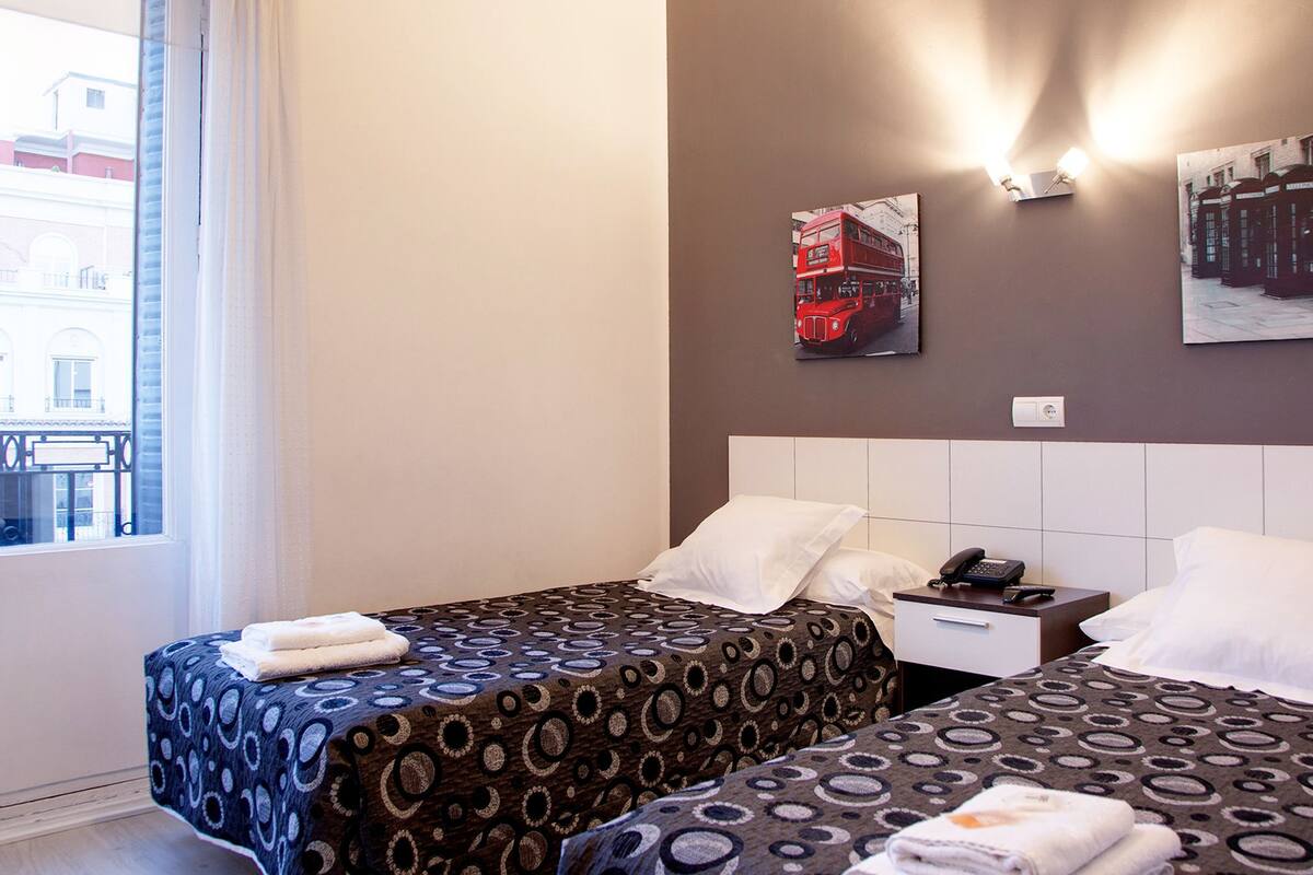 A hotel room with two beds, a nightstand with a phone, and a window with a view of buildings.