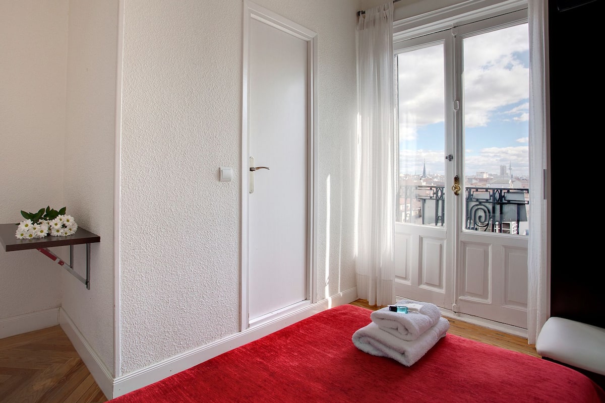 A bedroom with a red carpet, a white door, and a view of a cityscape through a large window.