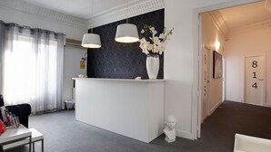 A hotel reception area with a white reception desk, two pendant lights, a vase with white flowers, a bust sculpture, and a hallway with a door marked 814. - Hostal Besaya (Madrid)