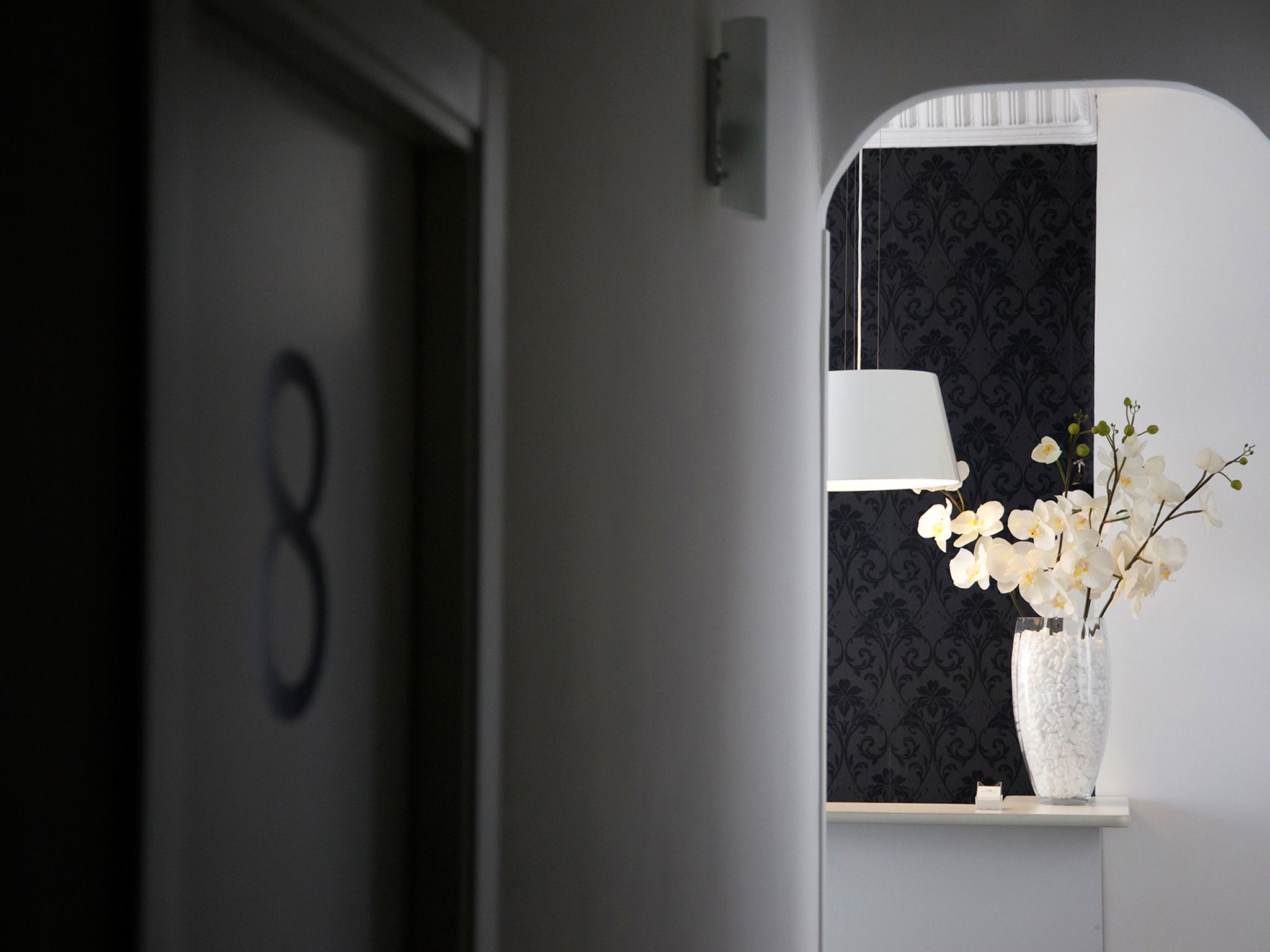A hotel room with a number '8' on the wall, a mirror with a white lampshade, and a vase with white flowers.