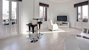 A modern living room with a large TV, a dining area with bar stools, and a view of the city through large windows. - Hostal Besaya (Madrid)