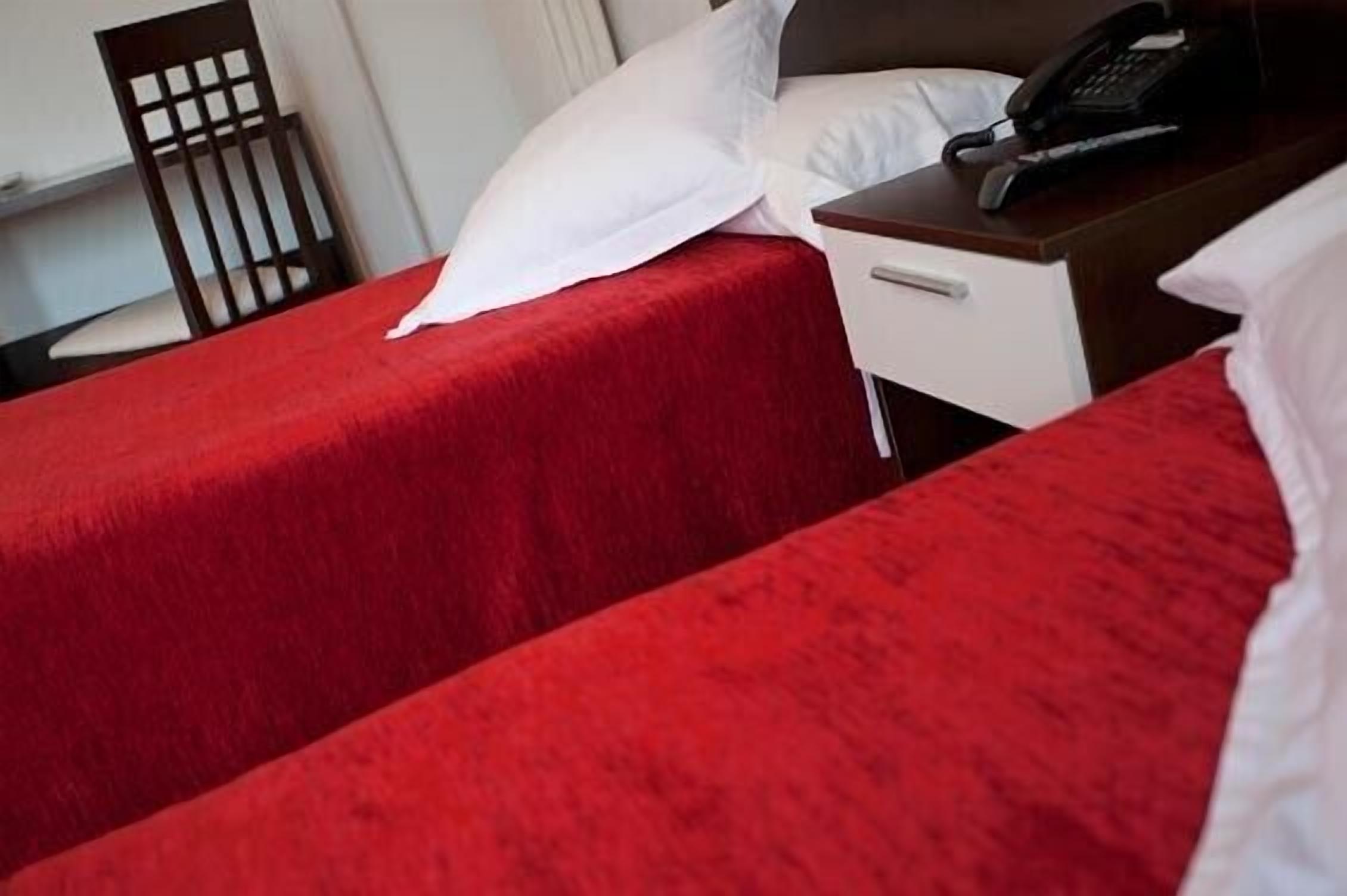 Ein Hotelzimmer mit zwei Betten, jeweils mit roter Bettdecke und weißen Kissen. Ein Holzstuhl und ein kleiner Tisch mit Telefon sind ebenfalls vorhanden.