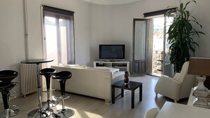 A modern living room with a sofa, two black bar stools, a TV, a dining table, and a potted plant. - Hostal Besaya (Madrid)