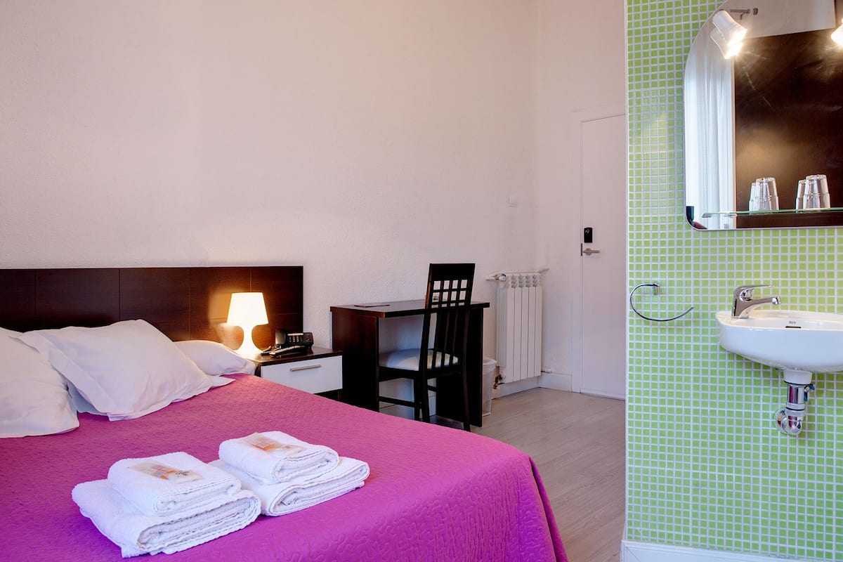 A hotel room with a bed, a desk, a chair, a lamp, a telephone, a radiator, and a bathroom with a sink and mirror.