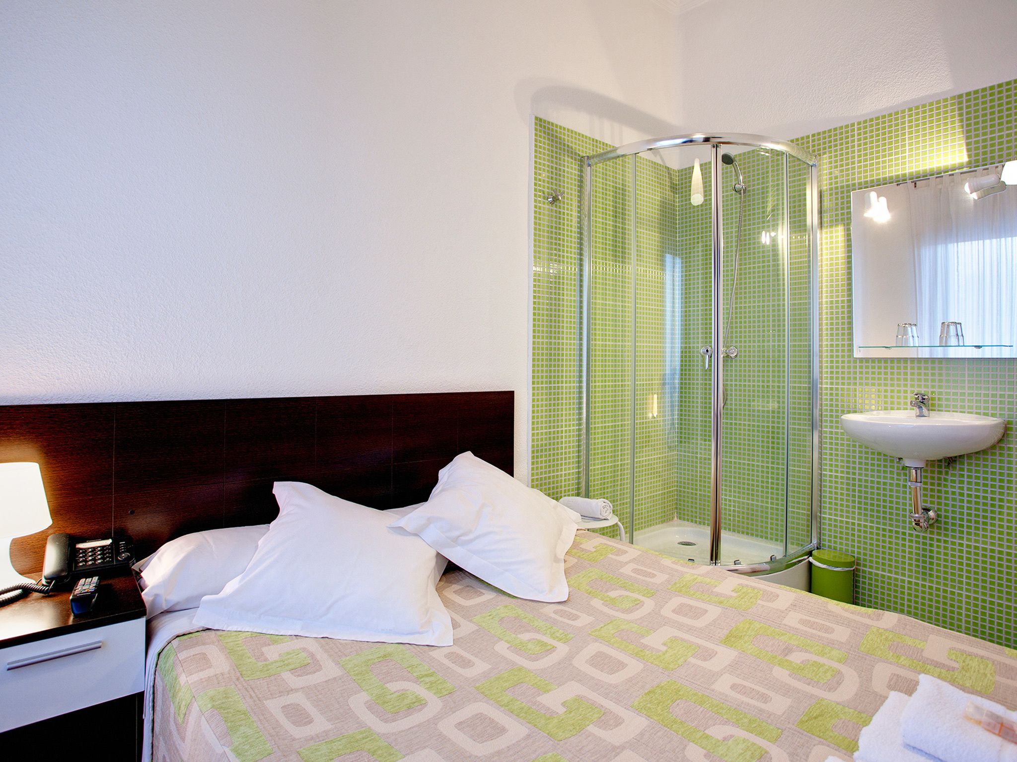 A hotel room with a bed, a bedside telephone, a green-tiled shower enclosure, and a sink with a mirror above it.