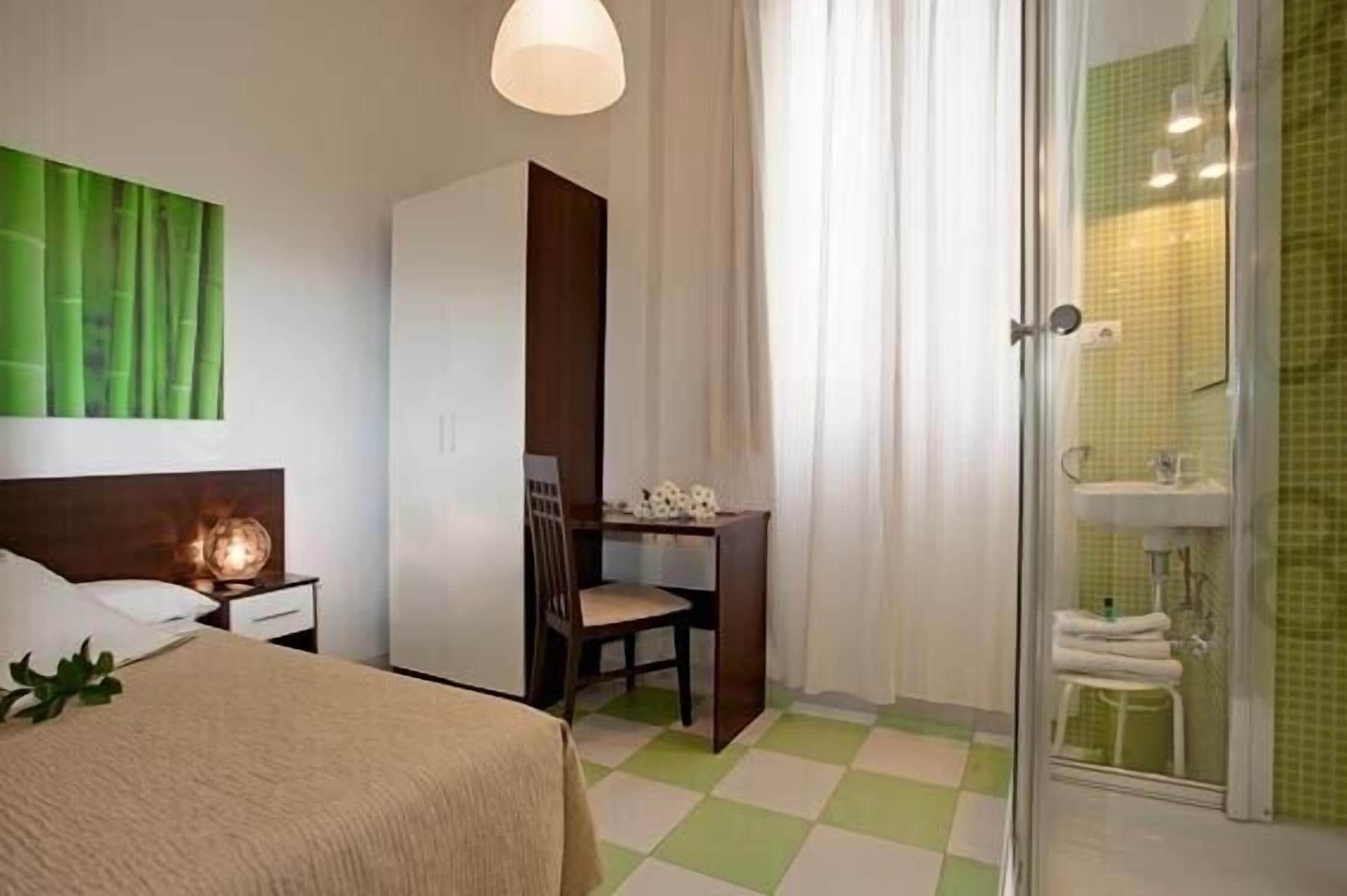 A hotel room with a bed, a desk, a chair, a shower area, and a green checkered floor.
