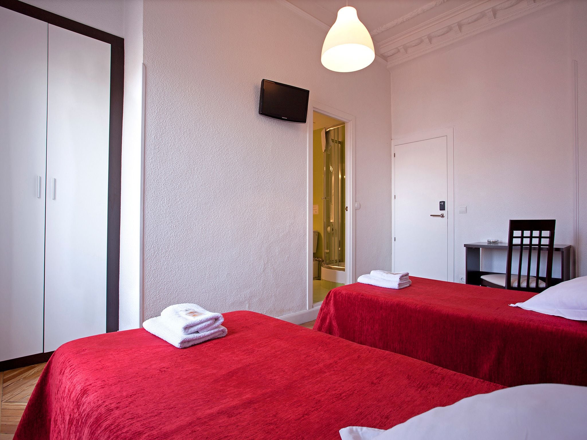 A hotel room with two single beds, a desk with a chair, a TV mounted on the wall, and a shower area visible through an open door.