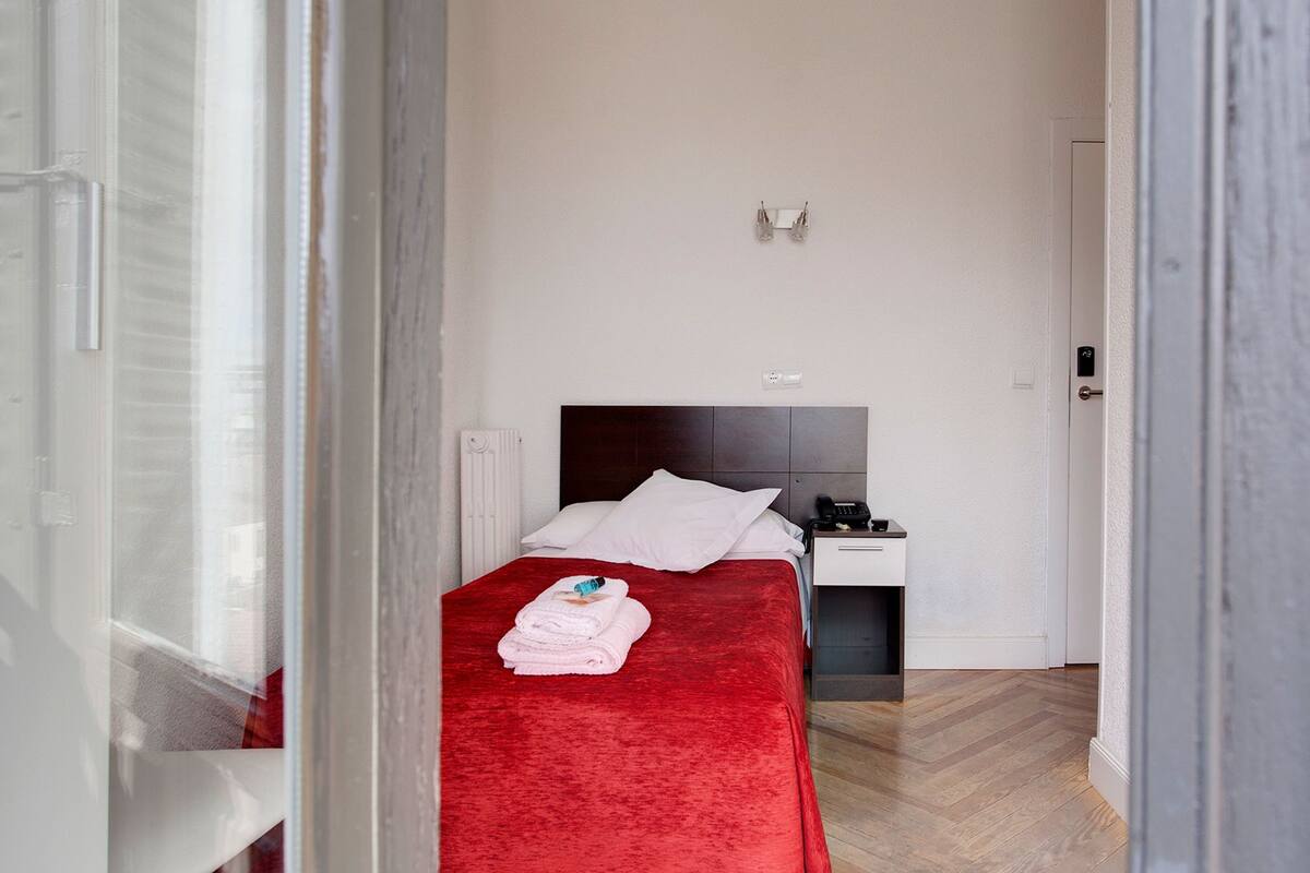 A hotel room with a red bedspread, white pillows, a bedside table with a phone, and a radiator.