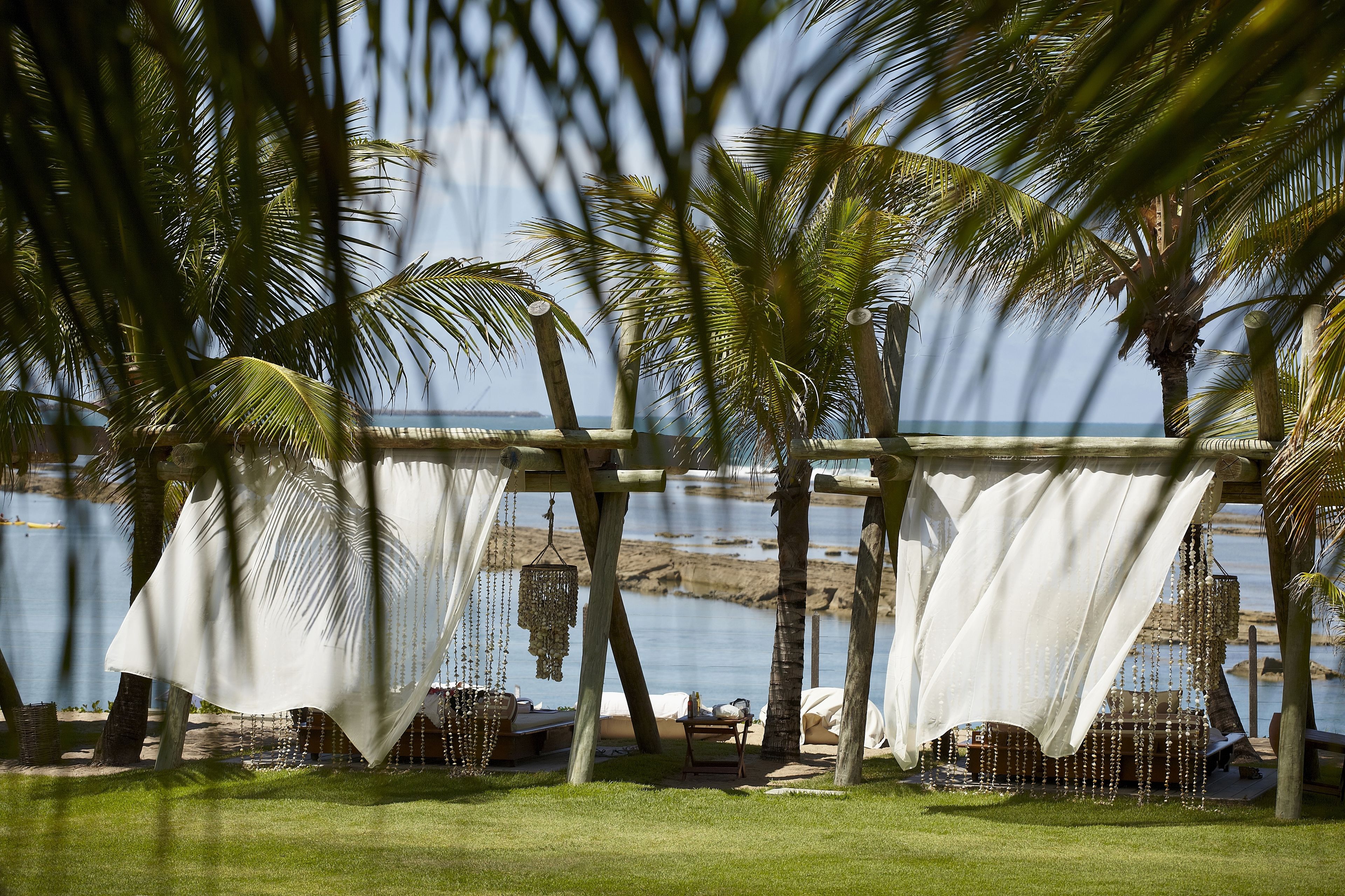 Um espaço de descanso à beira-mar com vista para o mar, com pérgola de madeira, cortinas brancas e cadeiras de lounge.