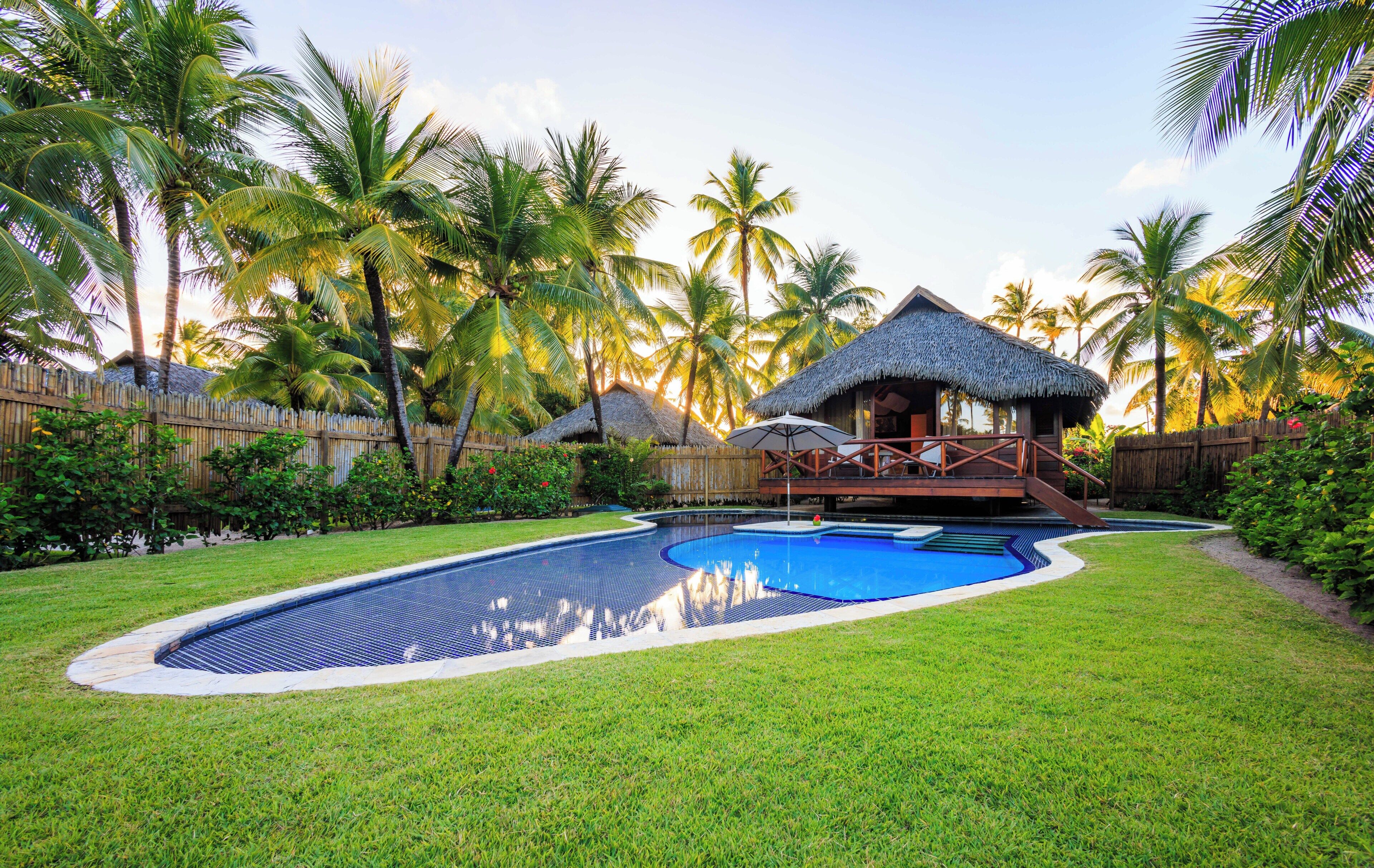 A swimming pool with a thatched-roof structure and a wooden deck in a tropical setting.